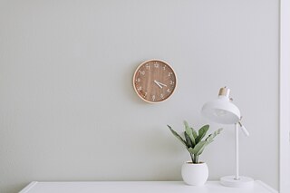 Vor einer weißen Wand steht ein weißer Tisch, darauf stehen eine weiße Lampe und eine Pflanze in einem weißen Übertopf. An der Wand hängt eine Uhr.