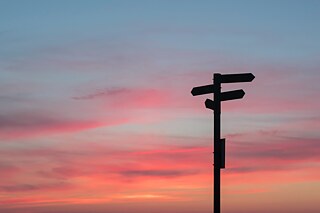 Ein Wegweiser im Gegenlicht fotografiert, im Hintergrund das Abendrot am Himmel.