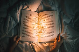 Ein Buch liegt aufgeschlagen auf einer Bettdecke. Darauf liegt eine Lichterkette.
