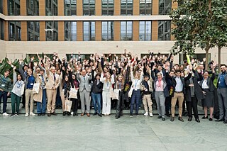 Gruppenfoto im Innenhof eines beigen Gebäudes mit Fensterfront. Alle Personen heben die Hände in die Luft und jubeln.