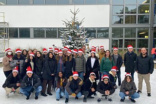 Gruppenfoto von Schülerinnen und Schülern mit roten Weihnachtsmützen vor einem Schulgebäude. Es liegt Schnee und im Hintergrund steht ein großer Tannenbaum