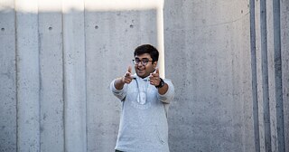 Junger Mann mit Brille und einem grauen Pullover zeigt mit beiden Händen Richtung Kamera und lacht