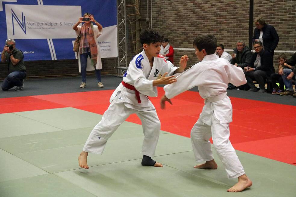 Zwei Jungen in weißen Judoanzügen beim Training auf Matten. Im Hintergrund zwei Personen, die fotografieren und sitzendes Publikum