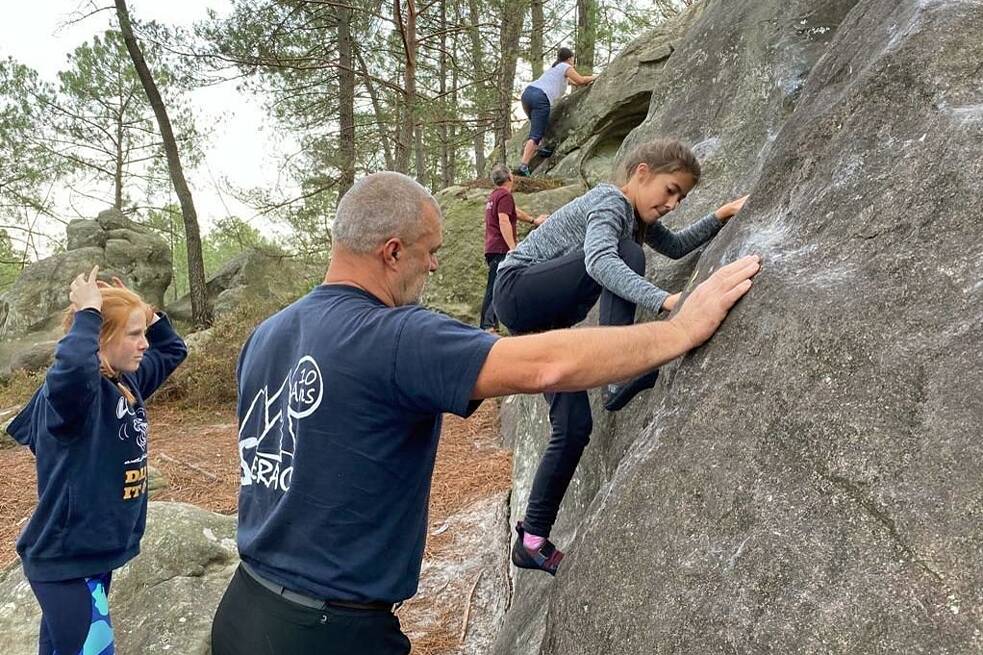 Mädchen klettert an einem Felsen hoch. Zwei weitere Personen stehen bei ihr. Im Hintergrund klettern weitere Personen.