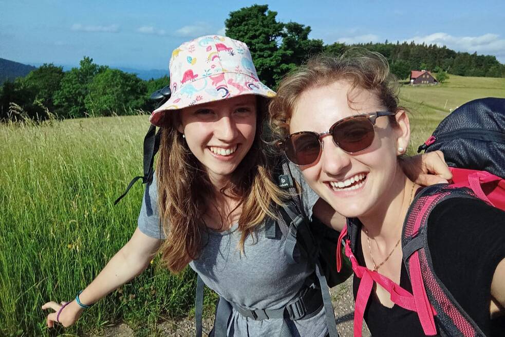 Selfie von zwei Jugendlichen mit Rucksäcken auf einer Wiese mit hohem Gras