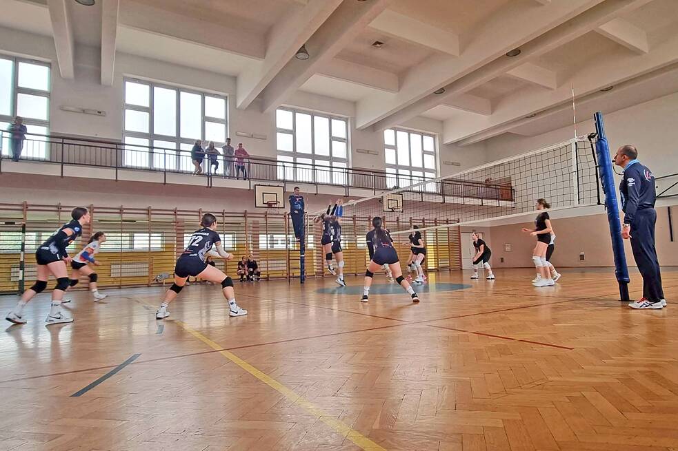 Mehrere Jugendliche spielen in einer Turnhallte Volleyball. Neben dem Netz steht ein Schiedsrichter, auf einer Ballustrade stehen Zuschauer und Zuschauerinnen