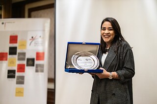 Eine Frau hält ein eingerahmtes Zertifikat in die Kamera, im Hintergrund ein Plakat der PASCH-Initiative