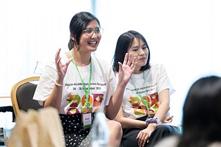 Zwei Frauen mit weißen T-Shirts, auf denen "PASCH-Alumni-Konferenz Bangkok" steht, sitzen nebeneinander. Die linke Frau spricht.