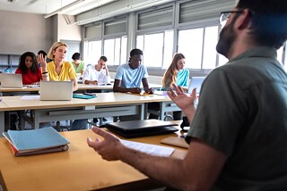 Ein Mann spricht zu Frauen und Männern, die vor ihm an Tischen in einem Klassenraum sitzen. Der Mann ist von der Seite fotografiert.