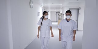 In Arbeitskleidung im Krankenhaus © privat Zwei Personen laufen in weißer Arbeitskleidung und FFP 2 - Masken über einen weißen Flur.