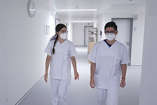 Zwei Personen laufen in weißer Arbeitskleidung und FFP 2 - Masken über einen weißen Flur.