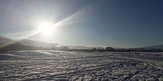 Eine schneebedeckte Landschaft im Sonnenschein, im Hintergrund sind ein paar Gebäude zu erkennen