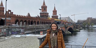 Larissa in einer braunen Jacke vor einer Brücke aus rotbraunen Steinen