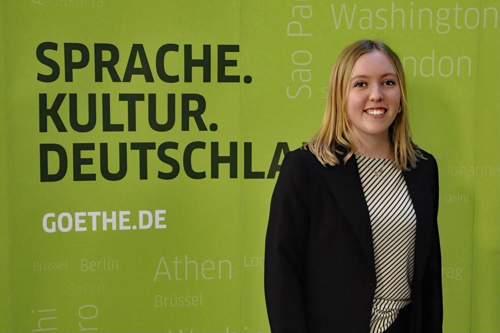 Eine blonde, junge Frau steht vor einem Plakat des Goethe-Instituts und lächelt in die Kamera.