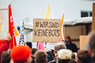 Eine Gruppe Menschen halten Schilder und Fahnen hoch. Auf einem Schild steht „#WIRSINDKEINEBOTS“.