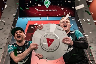 Zwei E-Sportler halten den Pokal der Clubmeisterschaft der Virtual Bundesliga und jubeln.