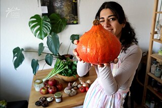 Eine junge Frau mit dunklen, langen Haaren steht in einer Küche und hält einen Kürbis in die Kamera. Sie trägt eine Schürze. Im Hintergrund sieht man einen Tisch und ein Regal bestückt mit Lebensmitteln, frisch und eingekocht.