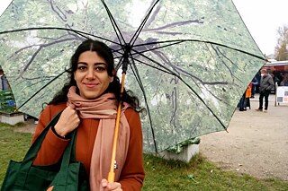 Eine junge Frau mit dunklen, langen Haaren steht unter einem großen Regenschirm auf einem Bauernmarkt.