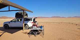 Ein Mann sitzt an einem Klapptisch, vor ihm stehen zwei Laptops. Im Hintergrund ein Geländewagen mit Zelt auf dem Dach und eine Wüstenlandschaft.