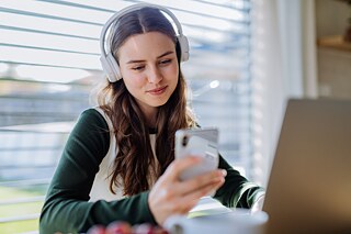 Eine junge Frau mit langen, braunen Haaren sitzt vor einem Laptop, blick auf ein Smartphone und trägt Kopfhörer. 