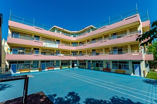 Pink-gelbes Schulgebäude mit bunten Bänken und Sportplatz im Vordergrund