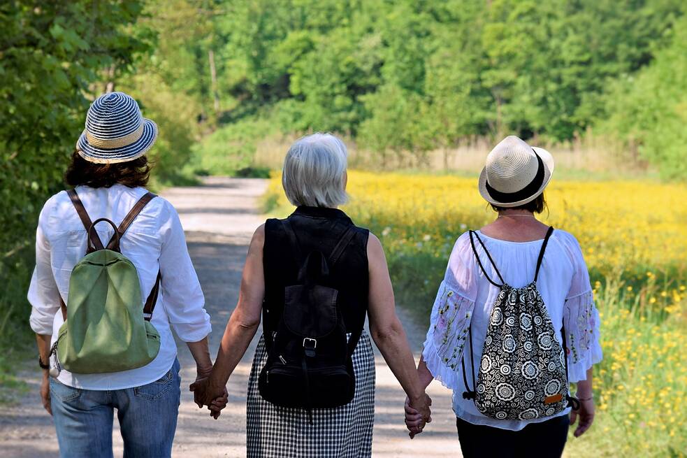 Drei Personen mit jeweils einem Rücksack auf dem Rücken gehen Hand in Hand spazieren. Zwei tragen Sommerhüte. 