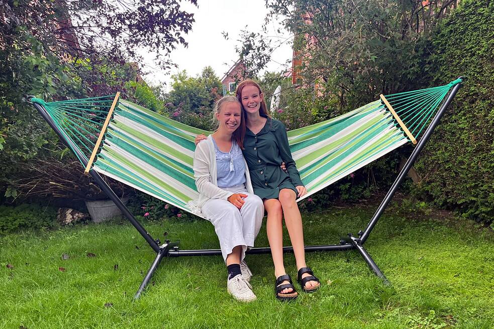 Zwei junge Frauen sitzen auf einer breiten Hängematte in einem Garten.