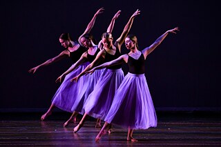 4 Schülerinnen in schwarzem Oberteil und lila Rock auf einer Bühne, tanzen in Formation, ein Bein vorgestellt, Arme gehoben