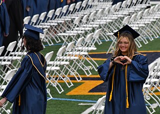 Zwei Schülerinnen in Graduation-Robe und Hut, im Hintergrund weiße Stuhlreihen auf Rasen; eine Schülerin lächelt in die Kamera und formt mit den Händen ein Herz