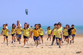 Kinder in gelben T-Shirts und blauen kurzen Hosen laufen fröhlich am Strand; im Hintergrund Meer und ein Kite-Surfer