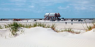 Im Vordergrund eine leicht bewachsene Sanddüne, im Hintergrund ein Pfahlbau am Strand von St. Peter Ording und dahinter das Meer.