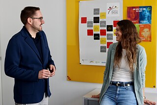 Die Verabredung Zwei junge Erwachsene stehen vor einer gelben Pinnwand mit Plakaten. Sie lächeln sich an.