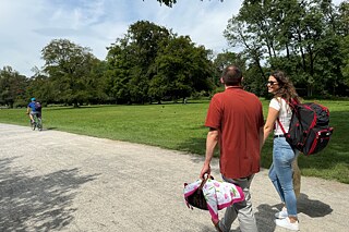 Auf dem Weg zum Biergarten Zwei junge Erwachsene auf einem befestigten Weg in einem Park. Sie trägt einen offenbar schweren, schwarz7roten Rucksack, er einen leichten Korb, dessen Inhalt mit einer Decke zugedeckt ist.