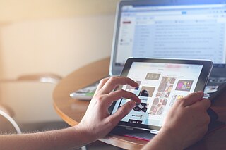 Zu sehen ist ein aufgeklappter Laptop auf einem Tisch stehend, davor ein Tablet, an dem die Hände eines Mannes zu sehen sind, die darauf tippen.