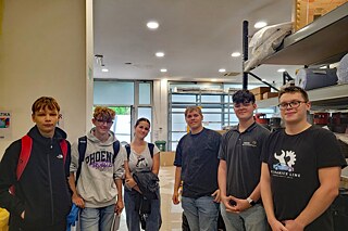 Sechs junge Menschen posieren für ein Foto, im Hintergrund sieht man hohe Regale und eine große Fensterfront.