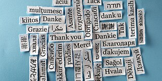 Auf einem hellblauen Hintergrund liegen weiße Zettel mit „Danke“ in unterschiedlichen Sprachen.