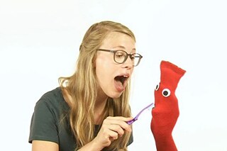 Eine blonde Frau putzt einer Handpuppe (rote Socke mit großen Kulleraugen) die Zähne.