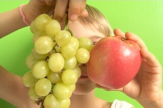 Ein kleines, blondes Mädchen hält einen roten Apfel und helle Trauben hoch.