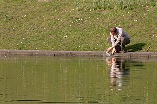 Ein etwa vierzigjähriger Mann steht an einem Weiher über das Wasser gebeugt und betrachtet sein Spiegelbild.