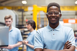 Ein junger Mann in einer Werkshalle. Er trägt eine Schutzbrille und ein hellblaues Polo-Shirt.
