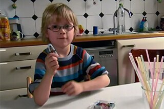 Ein kleiner Junge sitzt am Küchentisch. Er hält einen Strohhalm und eine Büroklammer in der Hand.