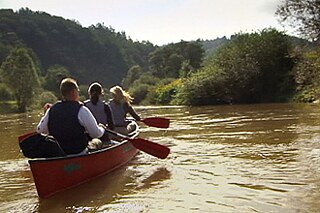 Auf einem kleinen Fluss paddeln drei Menschen in einem roten Boot.