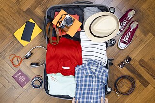Ein geöffneter Koffer liegt auf einem Parkettboden. Ein Pass, Kopfhörer, Sneaker, Sonnenbrille, Kleidung und anderes mehr liegen verstreut neben und im Koffer.