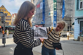Jugendliche auf einer Straße blicken auf ein s/w Foto eines alten Gebäudes.