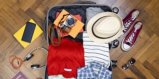Ein geöffneter Koffer liegt auf einem Parkettboden. Ein Pass, Kopfhörer, Sneaker, Sonnenbrille, Kleidung und anderes mehr liegen verstreut neben und im Koffer.
