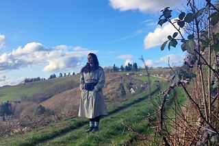 Eine junge Frau im hellen Regenmantel steht in einem Weinberg und blickt in die Ferne.