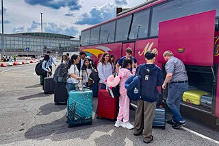 Jugendliche verladen zusammen mit einem kräftigen Erwachsenen Koffer in einen roten Reisebus.