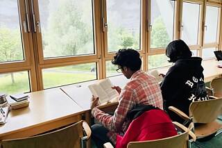 Zwei Jugendliche sitzen vor großen Fenstern an Tischen und halten Bücher in der Hand.