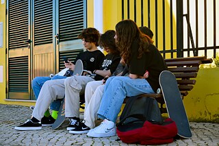 Jugendliche sitzen auf einer Bank und blicken auf ihre Smartphones. Neben ihnen stehen Scateboards und ein Rücksack.