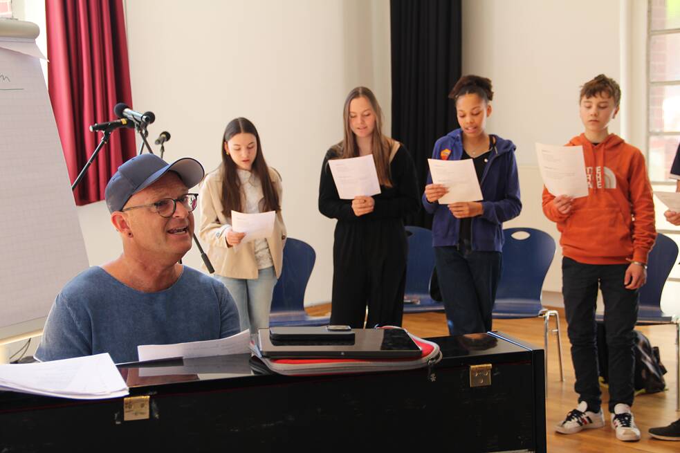 Vier Jugendliche halten Notenblätter in der Hand und singen; ein Erwachsener spielt auf einem Klavier.